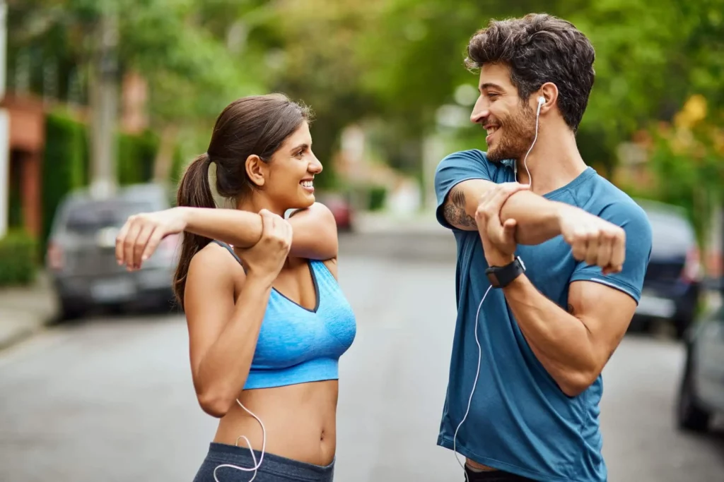 Imagem de uma mulher e um homem um de frente para o outro fazendo alongamento com roupa de academia no meio da rua cercados por carros e árvores para ilustrar matéria sobre as cidades futuristas