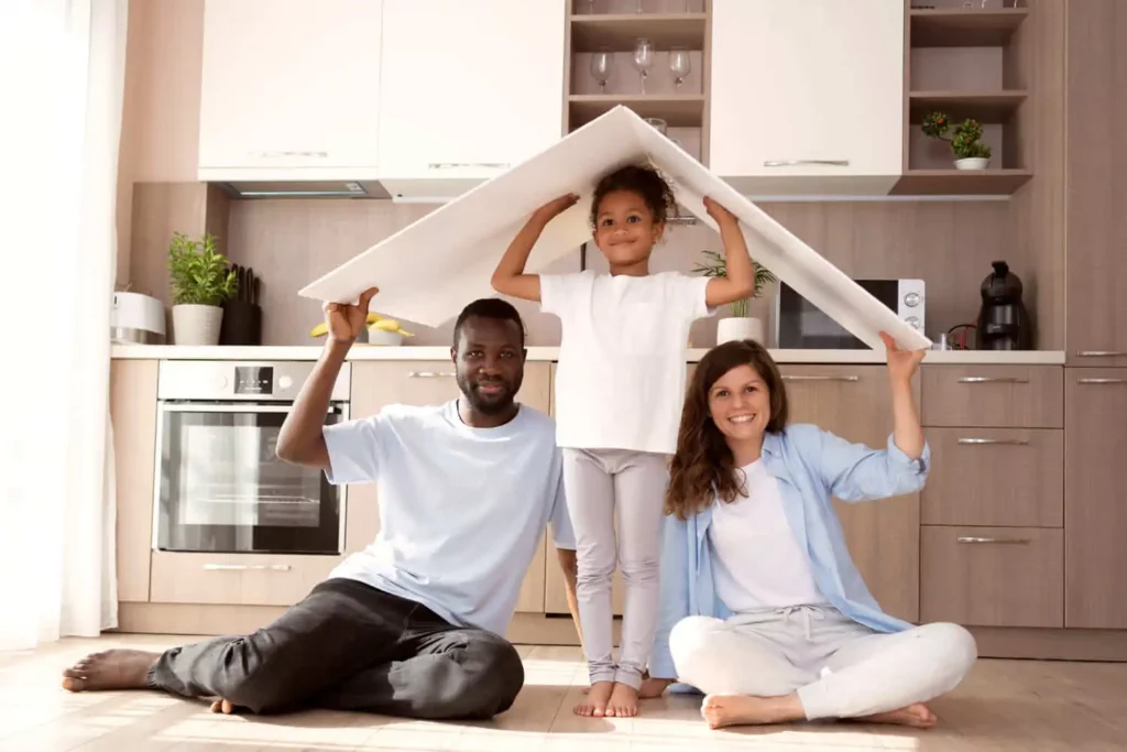 Imagem de uma família formada por um homem, mulher e criança sentados no chão segurando um objeto que lembra um telhado de casa para ilustrar matéria sobre como comprar apartamento sem entrada