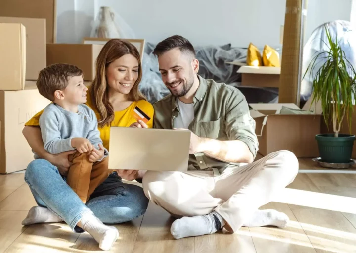 Imagem de uma família composta por um homem, uma mulher e uma criança sentados no chão de uma casa ao lado de caixas de papelão de mudança para ilustrar matéria sobre como comprar uma casa sem entrada