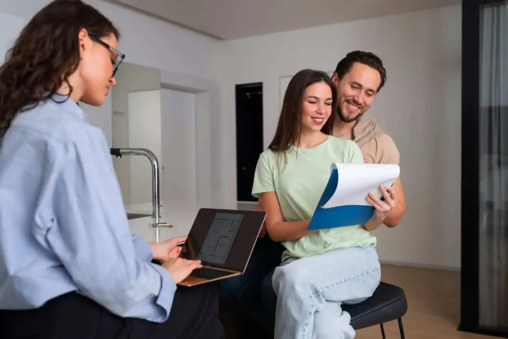 Imagem de um casal formado por um homem e uma mulher analisando um documento em frente a uma agente imobiliária que está com um notebook no colo para ilustrar matéria sobre como fazer uma proposta de compra de imóvel