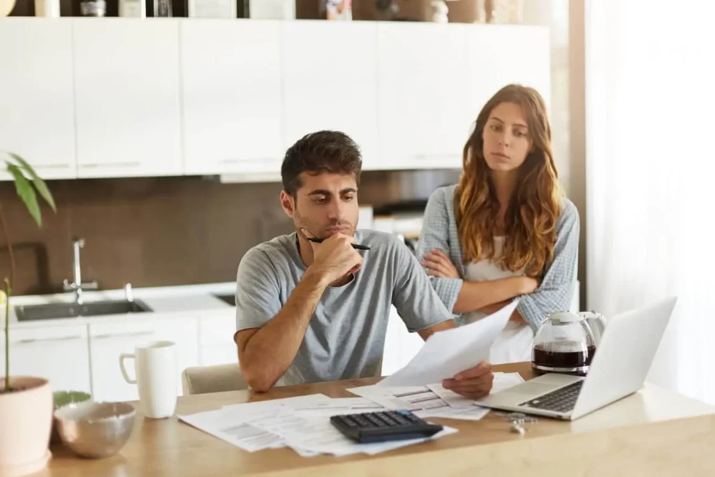 Imagem de um homem sentado em uma mesa de cozinha com expressão de preocupado enquanto segura papéis, com uma mulher atrás dele de braços cruzados, para ilustrar matéria sobre como comprar minha casa sem entrada. Eles estão cercados por documentos e um laptop.