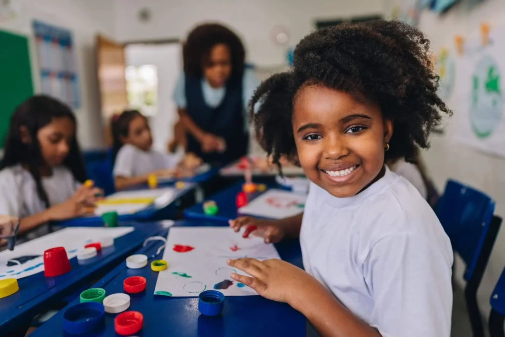  Imagem de uma criança sorridente em um colégio enquanto faz uma atividade de pintura em um papel que está em cima da carteira para ilustrar matéria sobre escola em Belford Roxo