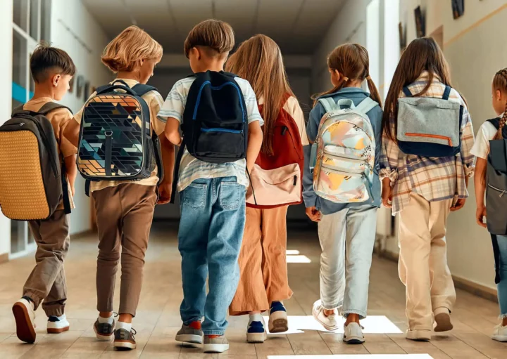 Imagem de crianças uma ao lado das outras de costas caminhando com mochila nas costas no corredor de uma instituição de ensino para ilustrar matéria sobre as escolas em Duque de Caxias