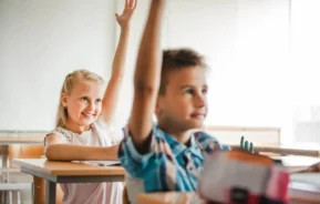 Imagem de um menino e uma menina sentados em carteiras em uma sala de aula olhando para a frente com os braços levantados chamando a professora para ilustrar matéria sobre as escolas em Betim