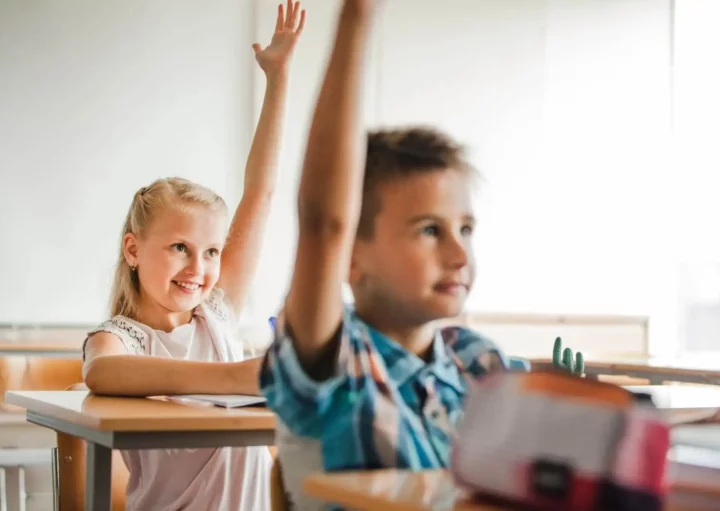 Imagem de um menino e uma menina sentados em carteiras em uma sala de aula olhando para a frente com os braços levantados chamando a professora para ilustrar matéria sobre as escolas em Betim