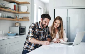 Imagem de um casal formado por um homem e uma mulher em pé na cozinha de um imóvel fazendo contas na bancada em frente a um computador para ilustrar matéria sobre a melhor data para amortizar financiamento