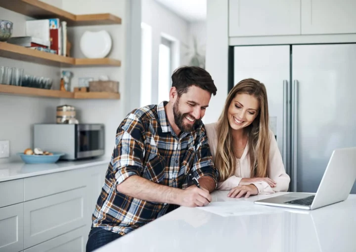 Imagem de um casal formado por um homem e uma mulher em pé na cozinha de um imóvel fazendo contas na bancada em frente a um computador para ilustrar matéria sobre a melhor data para amortizar financiamento