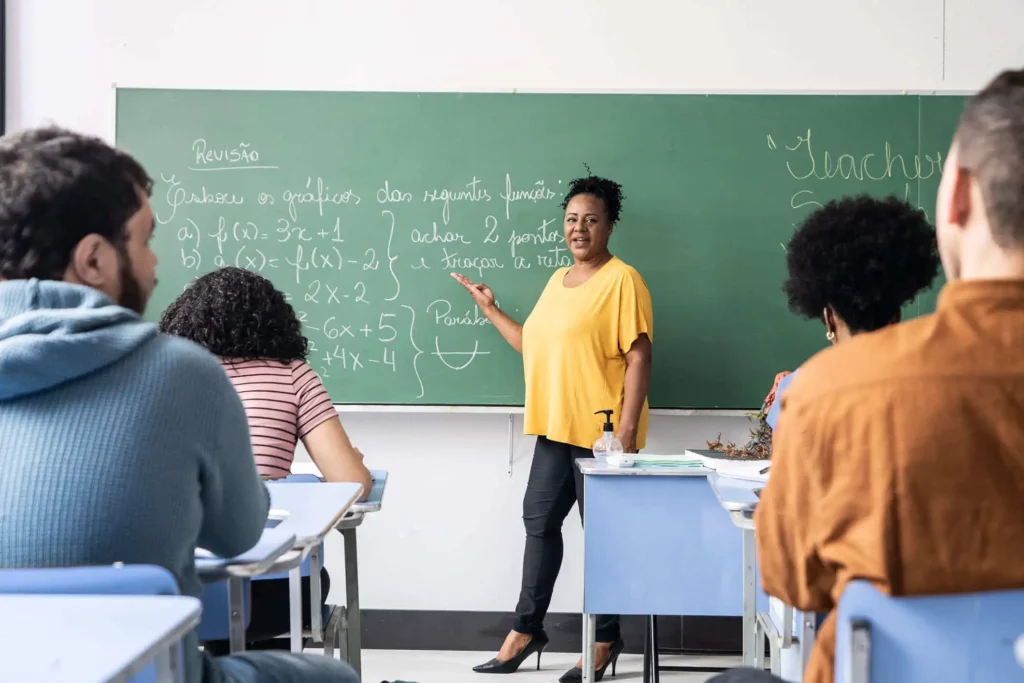 Imagem de uma turma de ensino médio onde os alunos estão sentados carteiras alinhadas assistindo a aula de uma professora que está em pé em frente ao quadro para ilustrar matéria sobre as melhores escolas em Belford Roxo