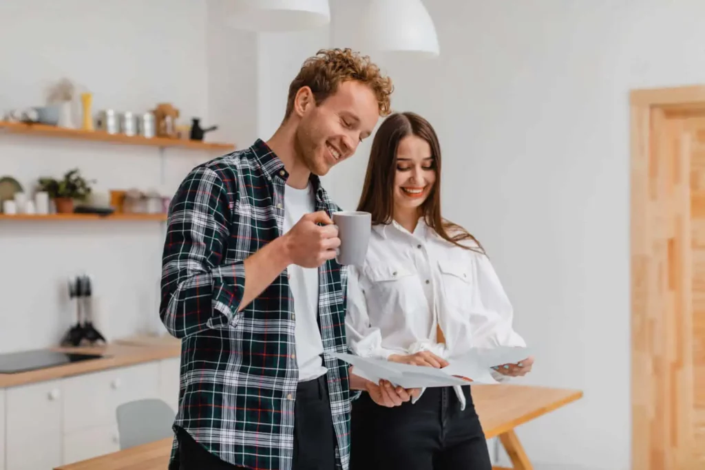 Imagem de um casal formado por um homem e uma mulher em pé na cozinha de um imóvel olhando alguns documentos enquanto o homem toma café em uma caneca para ilustrar matéria sobre modelo de proposta de compra de imóvel