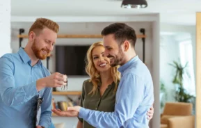 Imagem de um homem entregando um conjunto de chaves a um casal sorridente dentro de uma casa moderna e bem iluminada, simbolizando os novos proprietários que recebem as chaves após concluir o processo de inscrição imobiliária.
