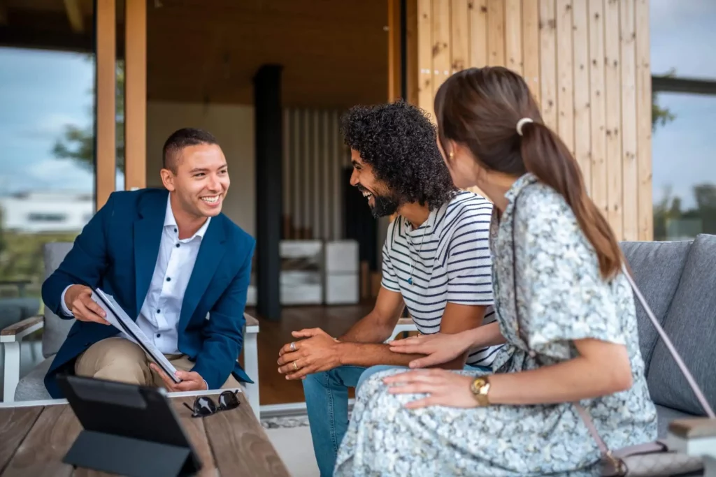 Imagem de um homem de blazer azul sorrindo enquanto mostra documentos a um casal casualmente vestido e sentado ao ar livre. O casal feliz está sentado em um banco próximo a uma construção de madeira.