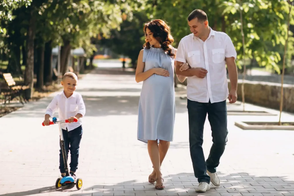 Imagem de uma família formada por um homem, uma mulher e uma criança para ilustrar matéria sobre o que fazer em São Paulo com crianças. Enquanto os pais estão caminhando de braços juntos, a criança está ao lado brincando de patinete na calçada de uma rua bem arborizada