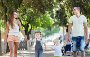 Imagem de uma família formada por um homem, uma mulher e duas crianças andando de mãos dadas na calçada de uma rua bem arborizada para ilustrar matéria sobre o que fazer em SP com crianças