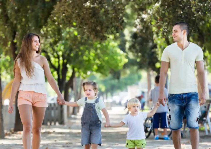 Imagem de uma família formada por um homem, uma mulher e duas crianças andando de mãos dadas na calçada de uma rua bem arborizada para ilustrar matéria sobre o que fazer em SP com crianças