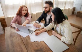 Imagem de um casal formado por um homem e uma mulher e uma terceira pessoa sentados ao redor de uma mesa de madeira olhando as informações contidas na tela de um notebook que está em cima da mesa ao lado de alguns papéis para ilustrar matéria sobre prenotação