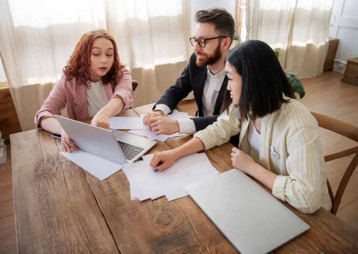 Imagem de um casal formado por um homem e uma mulher e uma terceira pessoa sentados ao redor de uma mesa de madeira olhando as informações contidas na tela de um notebook que está em cima da mesa ao lado de alguns papéis para ilustrar matéria sobre prenotação