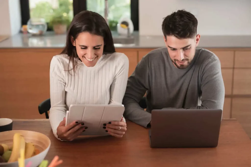 Imagem de um homem e uma mulher sentados lado a lado olhando as telas de um tablet e um notebook para ilustrar matéria sobre como fazer prenotação