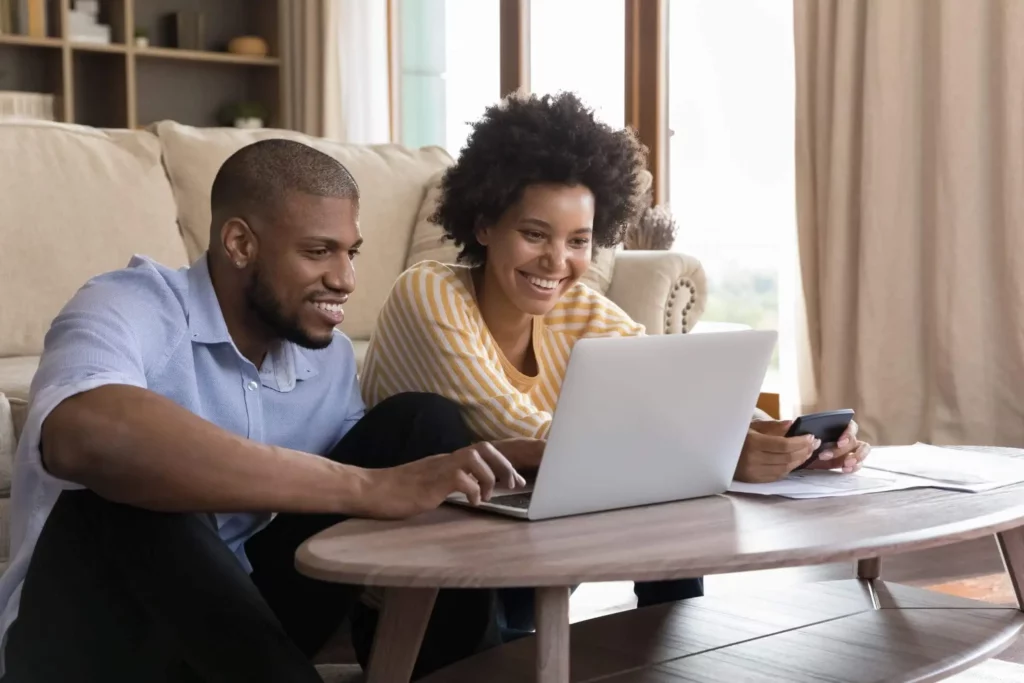 Imagem de um casal sentado no chão da sala de estar entre o sofá e uma mesa de centro olhando para a tela do notebook que está em cima da mesa de centro para ilustrar matéria sobre quando posso amortizar financiamento imobiliário