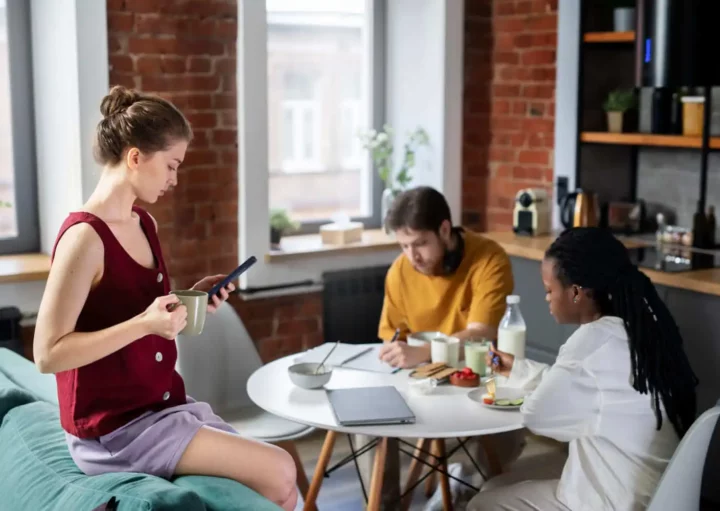 Imagem de uma mulher sentada na almofada de um sofá enquanto um homem e uma mulher estão sentados em frente em uma mesa com um notebook e vasilha em cima dentro de um imóvel para ilustrar matéria sobre se um herdeiro pode impedir a venda de um imóvel