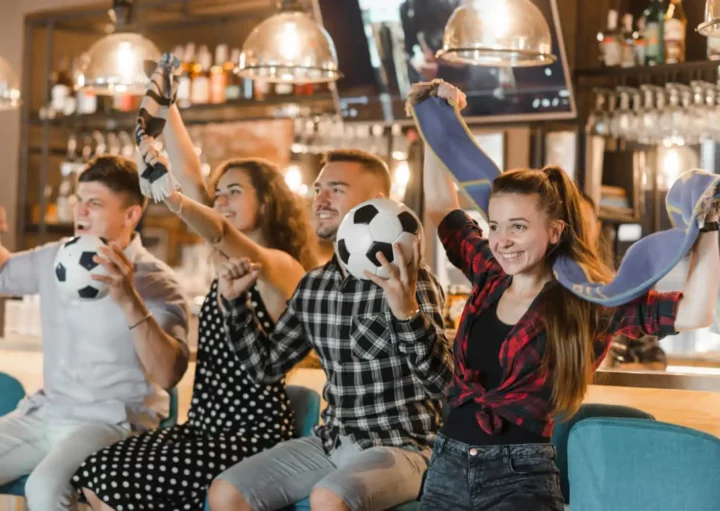 Imagem de dois homens e duas mulheres sentados no balcão de um bar torcendo com bolas de futebol na mão para ilustrar matéria sobre bar com telão