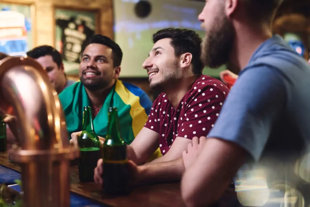 Imagem de um grupo de amigos em um bar com cervejas e bandeiras de times olhando fixamente para um ponto da imagem para ilustrar matéria sobre bar com telão para assistir jogo