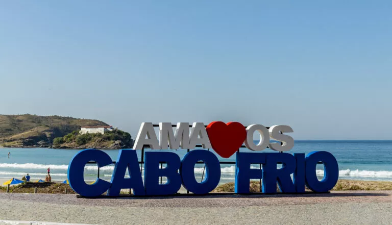 Imagem de um grande letreiro dizendo “Eu amo Cabo Frio” ao lado de uma praia de areia, com ondas e mar azul ao fundo em Cabo Frio, no Rio de Janeiro