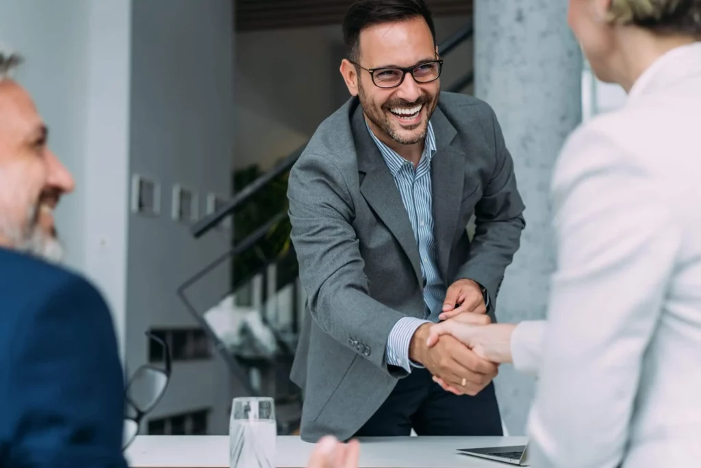  Imagem de um homem sorridente de terno apertando a mão de uma mulher que está acompanhada do marido para ilustrar matéria sobre a captação de imóveis direto com o proprietário