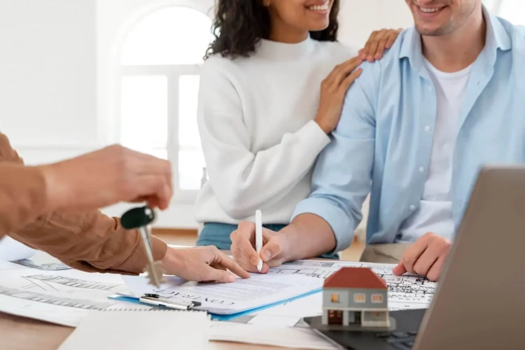 Imagem de um casal sorridente acenando documentação sobre uma mesa cheia de papéis e modelo de casa na presença de um agente imobiliário segurando uma chave para ilustrar matéria sobre como ter seu próprio apartamento