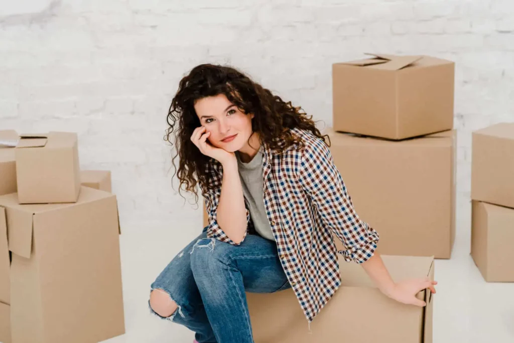 Imagem de uma mulher com meio sorriso sozinha centrada em uma caixa de mudança em um cômodo vazio para ilustrar matéria sobre se é possível financiar um apartamento sozinho 