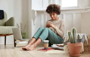 Imagem de uma mulher sentada no chão de casa ao lado de plantas, livros, um notebook, um copo de café e uma poltrona enquanto mexe no celular para ilustrar matéria que responde a pergunta: herdeiro pode ser expulso de imóvel?