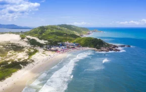 Imagem da vista aérea da Praia da Joaquina, em Florianópolis, mostra areia branca, ondas ondulantes e água azul clara, cercada por colinas verdes e penhascos rochosos sob um céu claro e ensolarado. Pessoas e prédios coloridos margeiam a vibrante orla