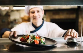 Imagem de um chef sorridente e uniformizado apresentando um prato preto com um prato gourmet colorido, guarnecido com verduras e legumes frescos, em um balcão de cozinha. Um sino de serviço pode ser visto nas proximidades.