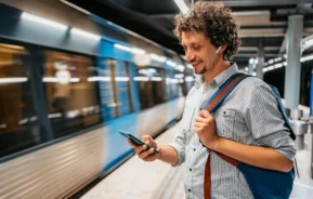 Imagem de um homem em pé em uma estação de metrô mexendo no celular enquanto um trem passa pela estação para ilustrar matéria sobre morar perto do metrô