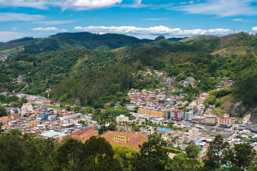 Imagem da vista panorâmica de Nova Friburgo, no interior do Rio, mostra colinas verdes, com prédios coloridos sob um céu azul brilhante e vegetação exuberante com árvores densas