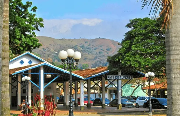 Imagem de uma praça com árvores, um poste de luz e um prédio marcado como Paty do Alferes. Carros estacionados e colinas verdes distantes são visíveis sob um céu azul