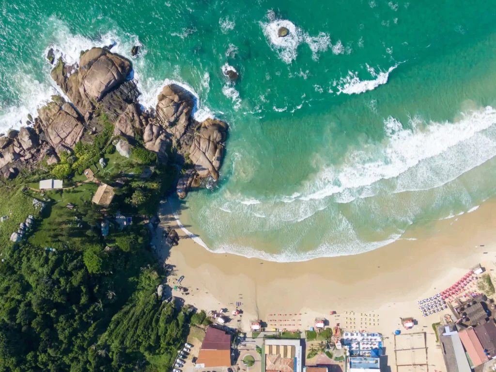 Imagem da vista aérea da Praia da Joaquina, em Florianópolis, mostra mar com ondas azul-turquesa, costa arenosa, penhascos rochosos à esquerda, vegetação exuberante e edifícios próximos à borda inferior. Guarda-sóis e pessoas são visíveis ao longo da orla