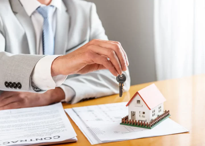 Imagem de um agente imobiliário com terno e gravata segurando a chave de um imóvel próximo a uma mesa com documentos para ilustrar matéria sobre recibo de aluguel
