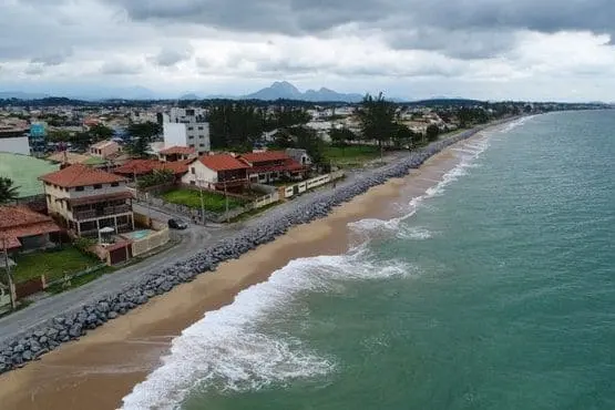 Imagem da vista aérea de Rio das Ostras, no interior do Rio de Janeiro, mostra casas perto da costa, uma barreira rochosa ao longo da praia, ondas batendo na areia e céu nublado acima
