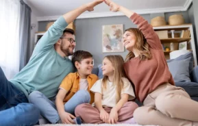 Imagem de uma família formado por um homem, uma mulher e duas crianças sentadas no sofá que está dentro de uma sala de um imóvel enquanto o homem e a mulher fazem o símbolo de um telhado com as mãos sobre as crianças para ilustrar matéria sobre o seguro DFI