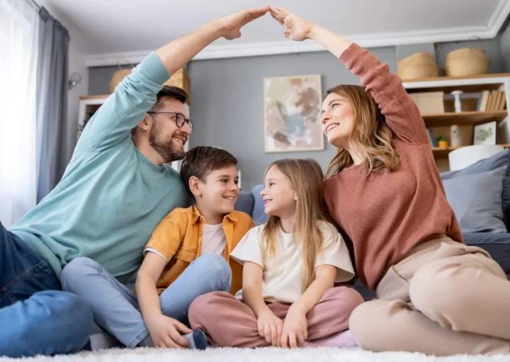 Imagem de uma família formado por um homem, uma mulher e duas crianças sentadas no sofá que está dentro de uma sala de um imóvel enquanto o homem e a mulher fazem o símbolo de um telhado com as mãos sobre as crianças para ilustrar matéria sobre o seguro DFI