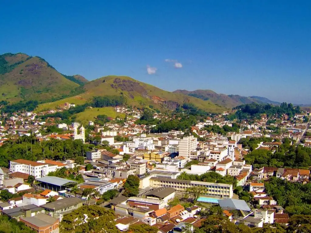 Imagem da vista aérea de Valença, uma das cidades do interior do Rio, mostra aglomerados de prédios e casas brancas, cercada por colinas e montanhas verdes sob um céu azul claro 