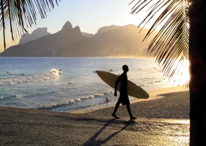 Foto que ilustra matéria sobre as Melhores praias para surfar no Brasil mostra a silhueta de um surfista com a prancha de baixo do braço com a praia do Arpoador ao fundo (Foto: Getty Images)