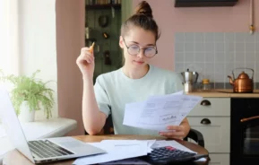 Imagem de uma mulher sentada ao redor de uma mesa de madeira na cozinha de casa analisando algumas contas para ilustrar matéria sobre contribuição predial. Enquanto ela segura algumas contas, outras estão em cima da mesa abaixo de uma calculadora e ao lado de um notebook