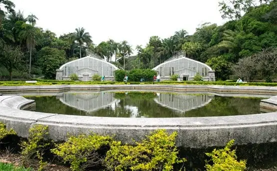Imagem do Jardim Botânico de São Paulo mostra um lago refletor circular com bordas baixas de pedra diante de duas estufas brancas, cercadas por uma vegetação exuberante.
