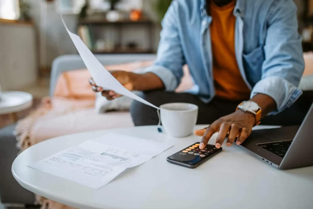 Imagem de um homem em casa sentado no sofá gerenciando as finanças ao fazer as contas na calculadora do celular para ilustrar matéria sobre o que é contribuição predial