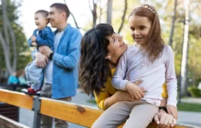 Imagem de uma mulher sorridente abraçando uma menina enquanto um homem segura um menino pequeno ao fundo. Aproveitando o que fazer nas férias em SP, eles relaxam ao ar livre em um parque iluminado pelo sol com árvores. Todos parecem felizes e despreocupados.