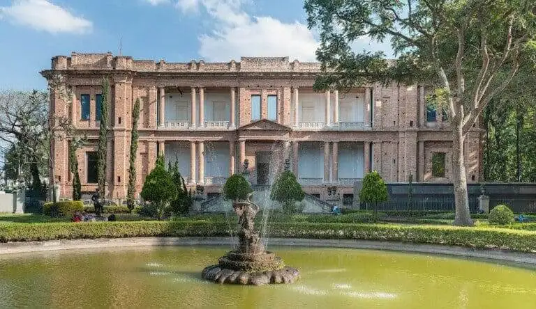 Imagem da Pinacoteca de São Paulo mostra um grande e histórico edifício de tijolos com colunas altas que fica atrás de uma fonte circular em um jardim paisagístico cercado por árvores e arbustos aparados sob um céu parcialmente nublado.