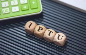 Imagem das iniciais “IPTU” escrita em um molde de madeira perto de uma calculadora verde para ilustrar matéria sobre quem paga o IPTU em caso de venda do imóvel