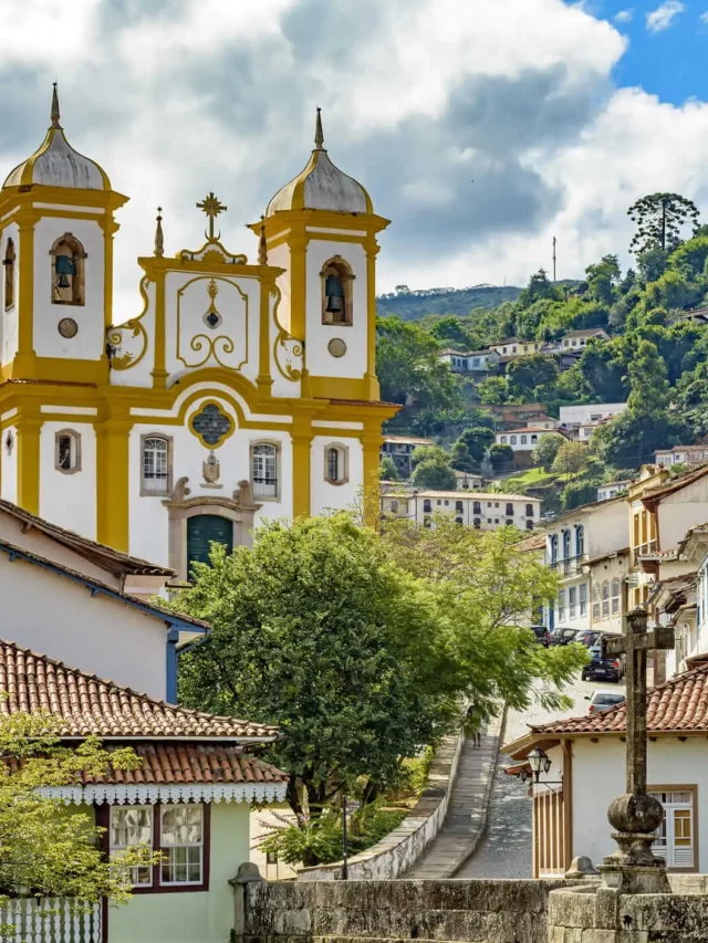 Uma igreja amarela e branca em estilo colonial com torres de sino gêmeas fica em uma rua de paralelepípedos, cercada por casas coloridas e colinas verdejantes sob um céu parcialmente nublado.