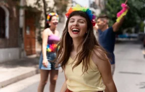 Imagem de mulher jovem sorridente e fantasiada para o carnaval de rua em primeiro plano enquanto dois amigos conversam em segundo plano para ilustrar matéria sobre as melhores cidades para curtir o carnaval